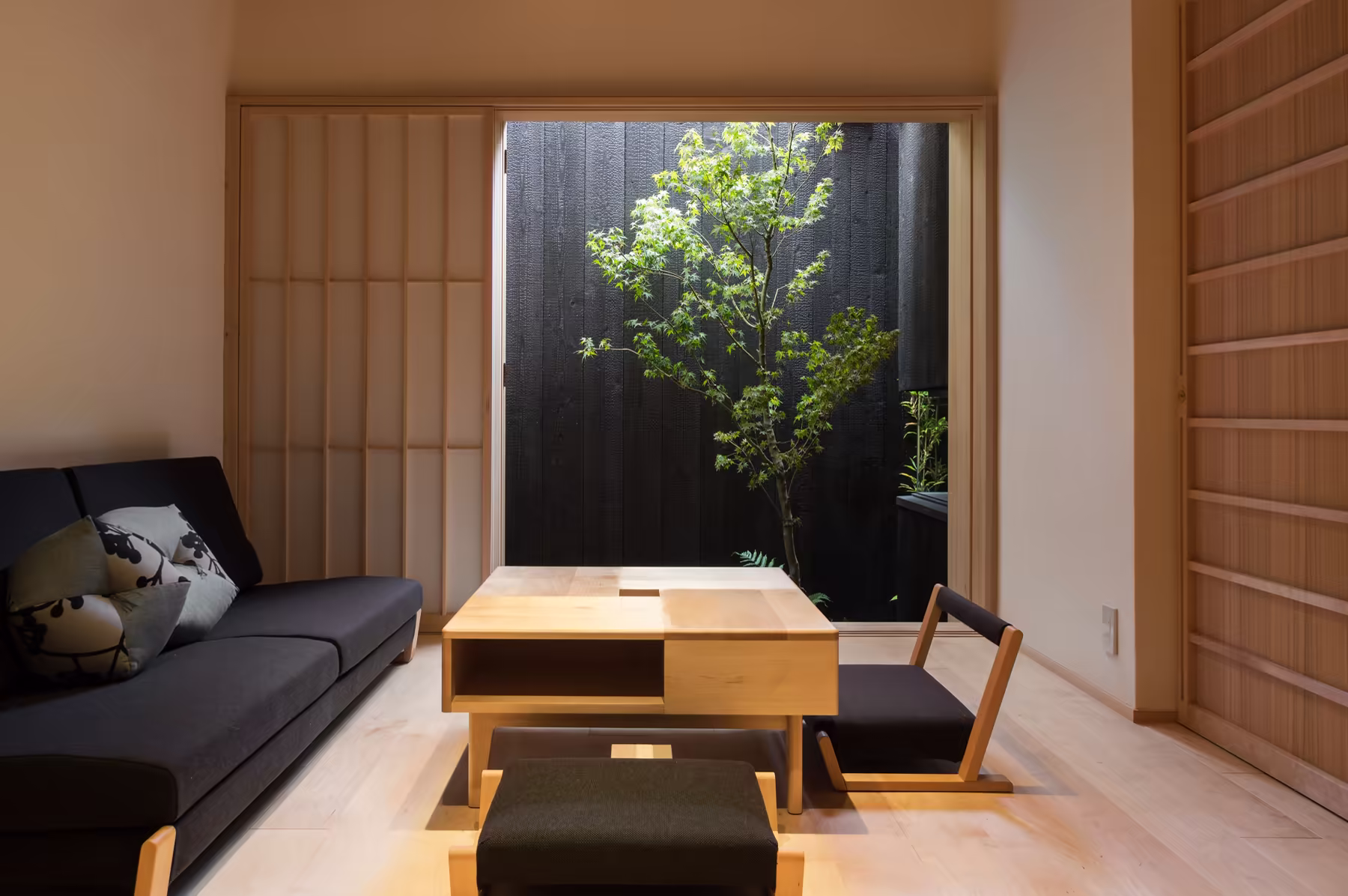 The living room — a long low sofa and a wooden table on the timber floor, with the courtyard maple framed in the opening beyond
