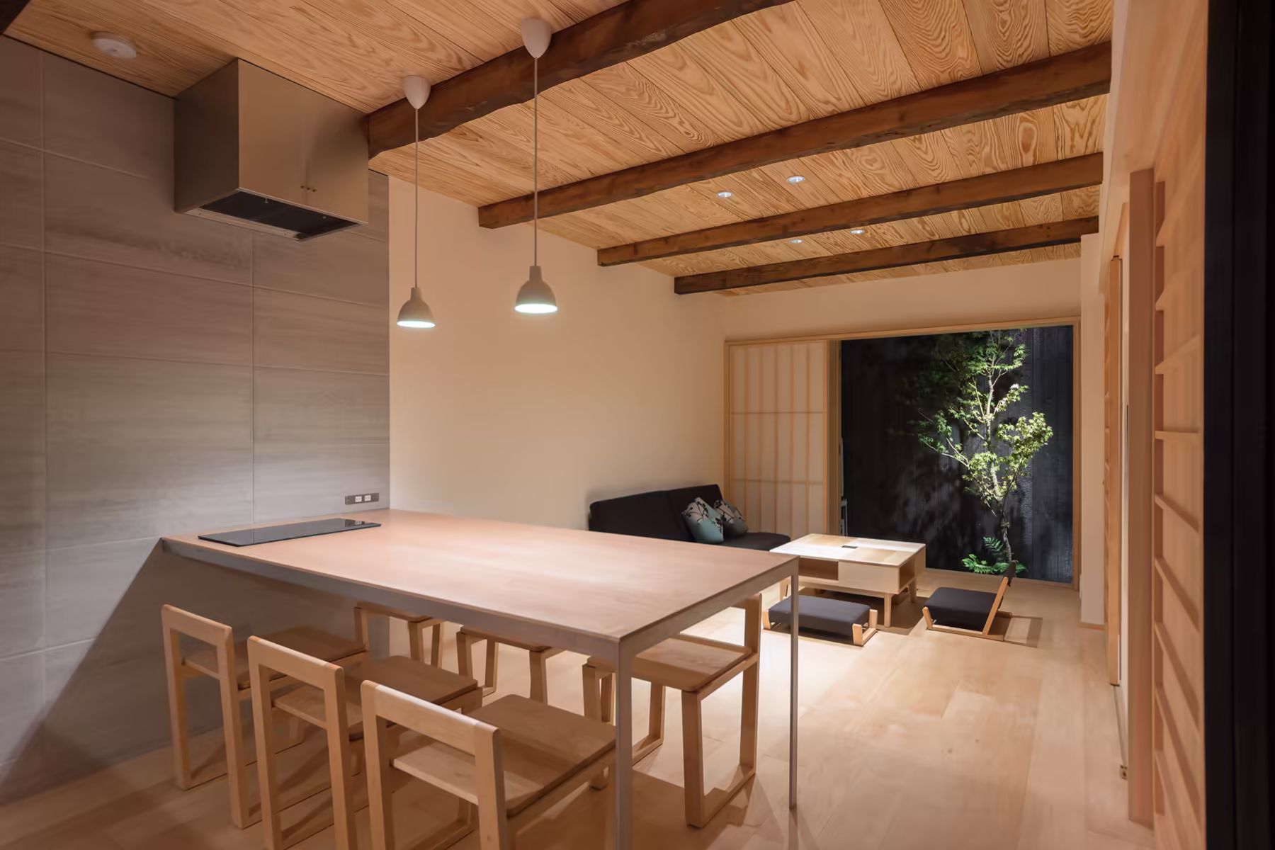 The lower storey of BenTen East — kitchen counter and dining table under the exposed timber ceiling, opening through to the living room and the courtyard maple beyond