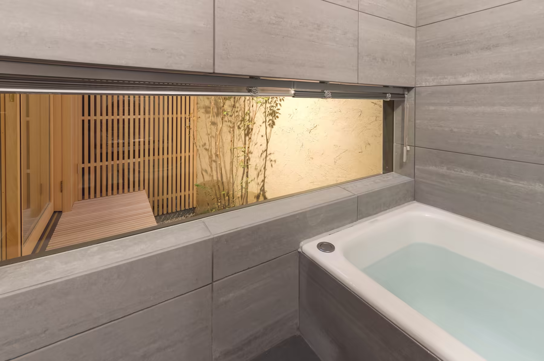 The bathroom — a deep tub in pale stone tile, with a long window onto the lit bamboo and slatted screen
