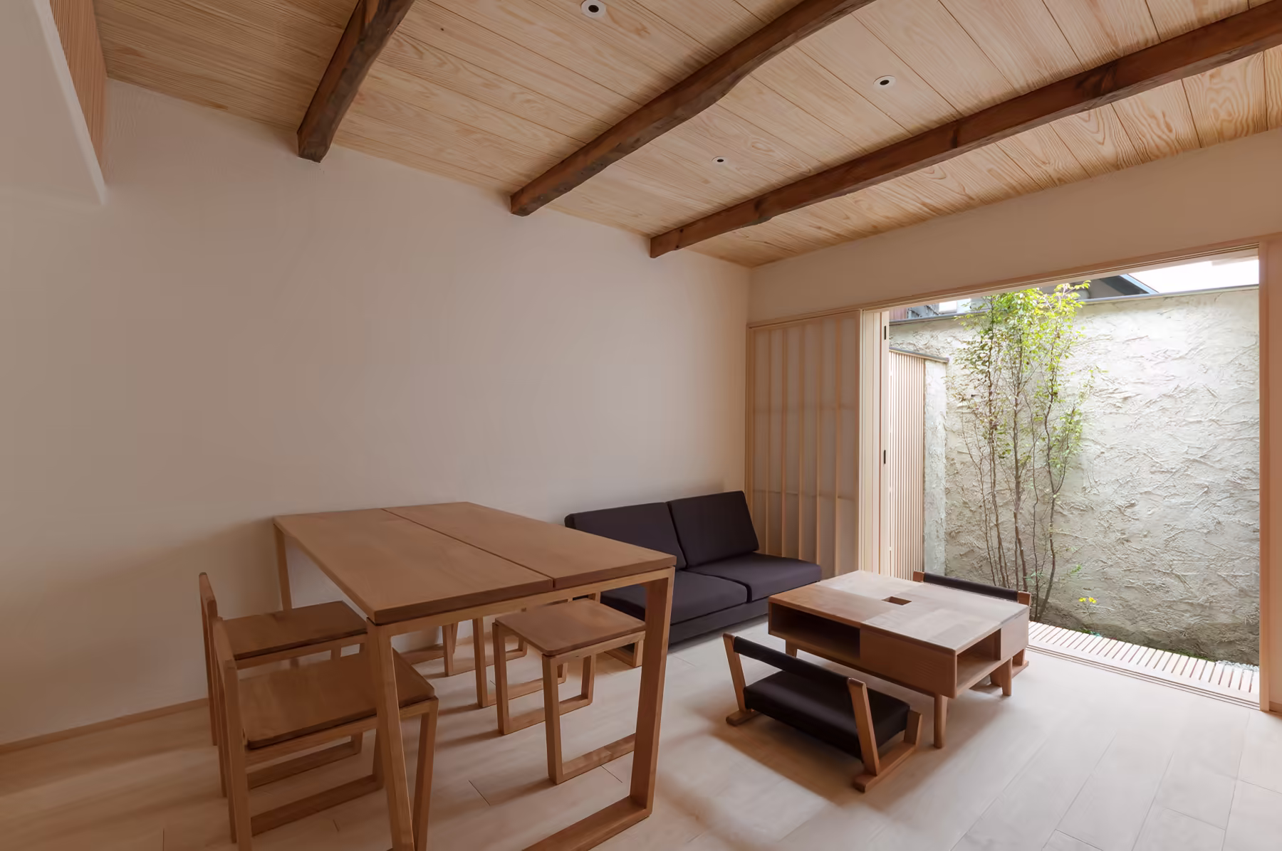 The dining table and chairs beside the long sofa, with the courtyard and its slender trees through the open wall